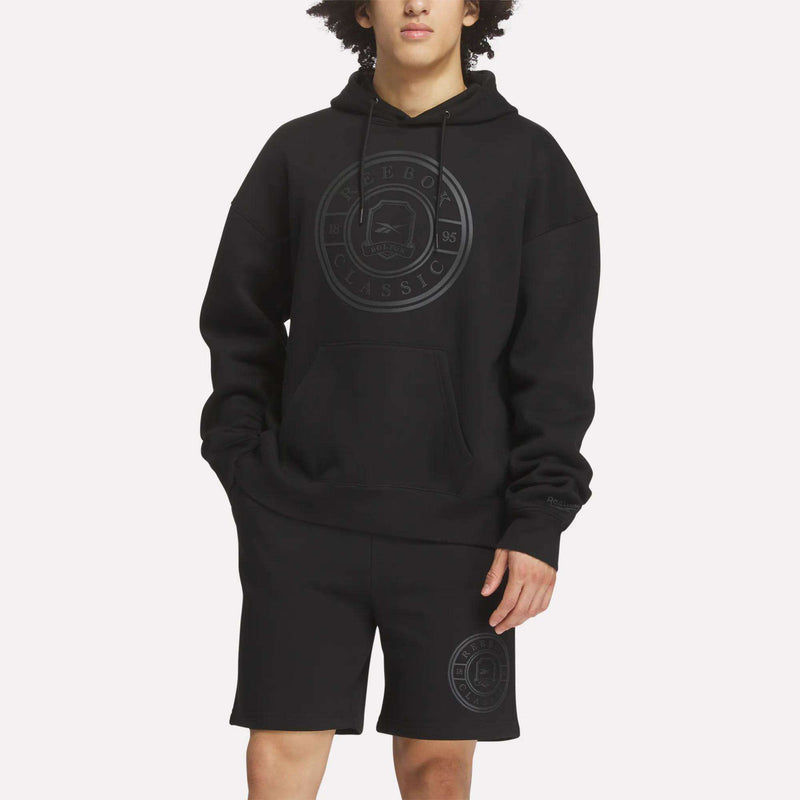 A person wears a relaxed-fit Men's Crest Hoodie by Reebok and black shorts, both with a subtle circular Reebok Classic logo, styled with hands in pockets against a plain light background.