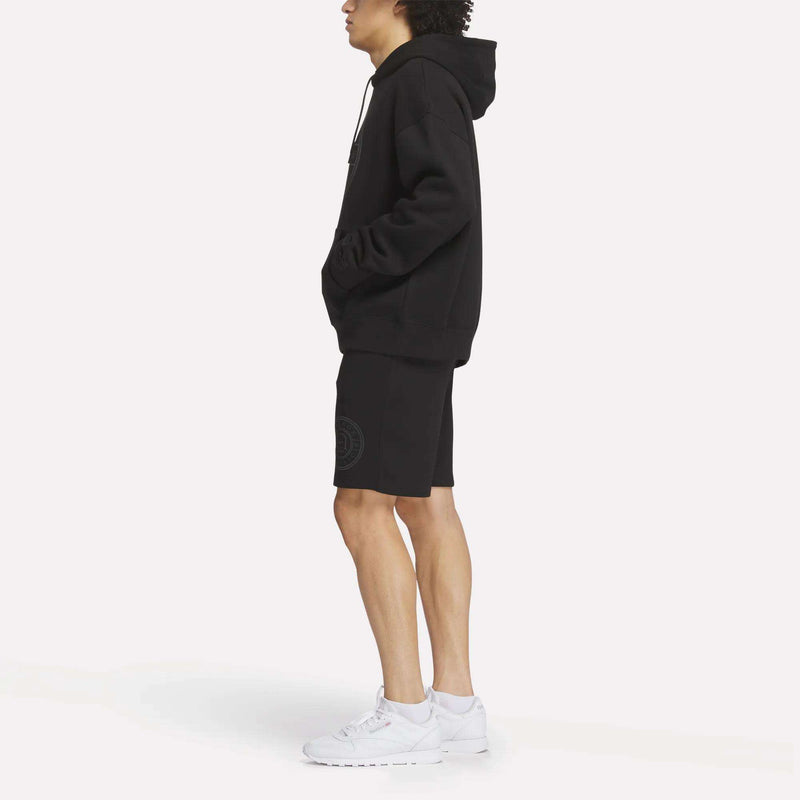 A person in profile wears the Reebok Men's Crest Hoodie, paired with black shorts and white sneakers, standing against a plain light background with hands in the hoodie pockets.