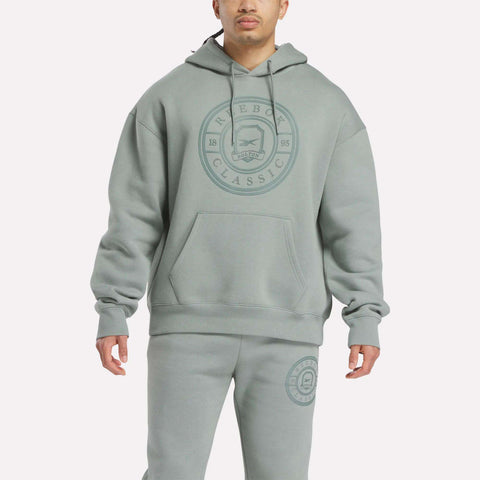 A person wears a relaxed fit, light gray-green Reebok Men's Crest Hoodie and matching fleece sweatpants with the circular logo. The hoodie features drawstrings and a front pocket. The background is plain white.