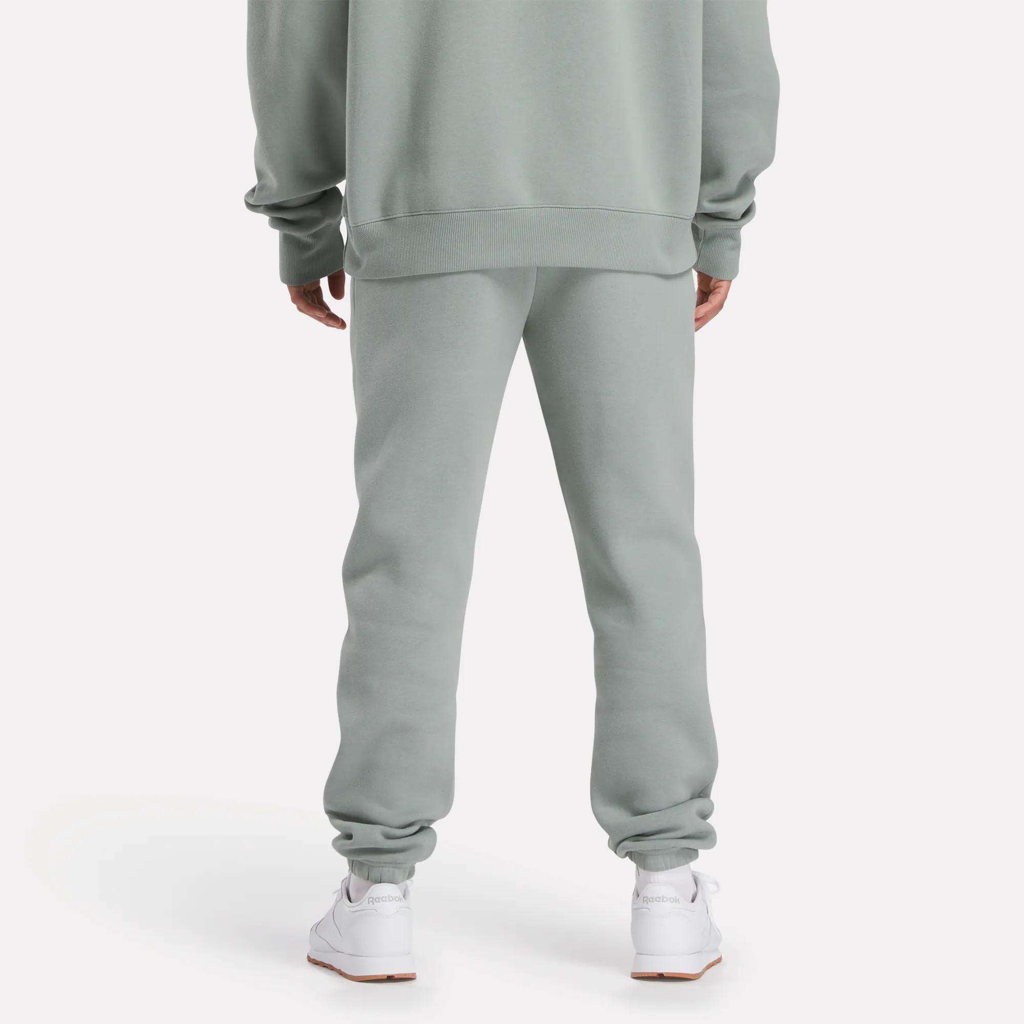 A person stands with their back to the camera, wearing light gray Reebok Men's Crest Jogger Pants and a matching sweatshirt for a casual look, paired with white sneakers against a plain light background.