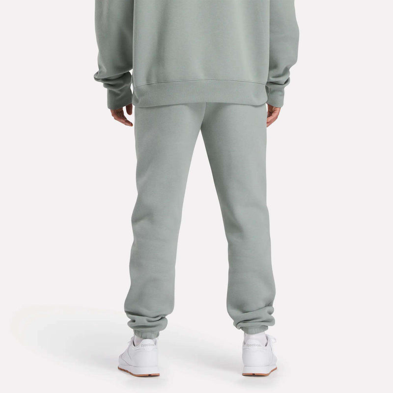 A person stands with their back to the camera, wearing light gray Reebok Men's Crest Jogger Pants and a matching sweatshirt for a casual look, paired with white sneakers against a plain light background.
