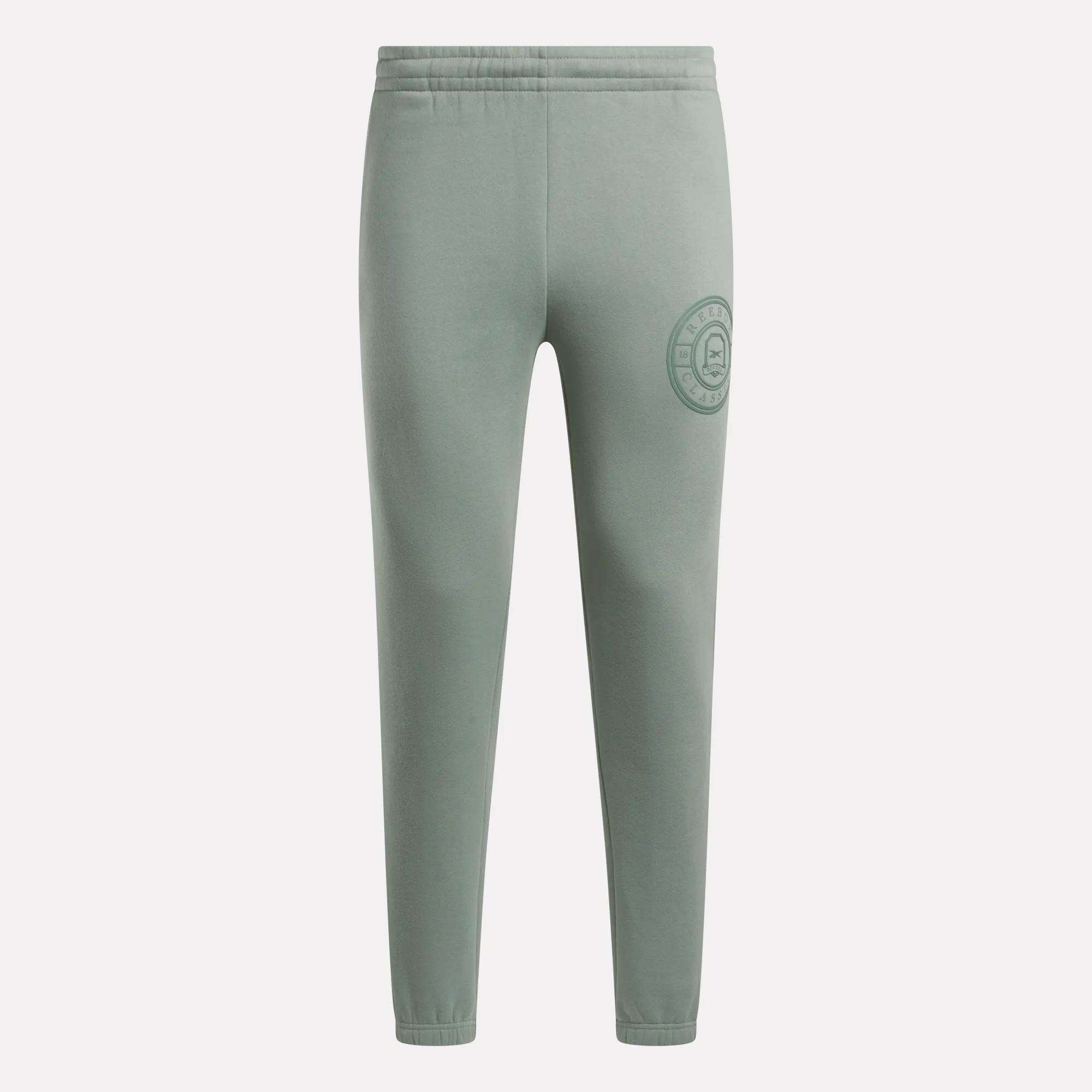 Light green Men’s Crest Jogger Pants by Reebok with an elastic waistband and a circular emblem on the upper left thigh. These cotton joggers feature a slim-fit design, offering casual comfort without visible pockets or drawstrings.