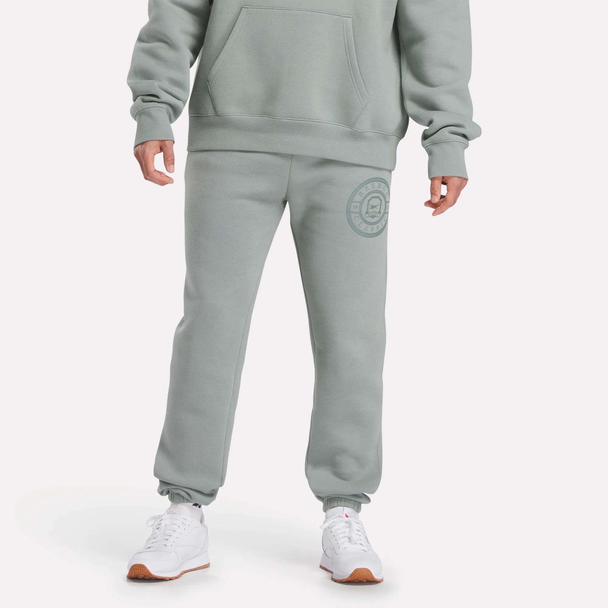 A person in light green Reebok Men's Crest Jogger Pants featuring a circular logo on the upper left thigh, paired with a matching sweatshirt and white athletic sneakers, stands against a plain white background.