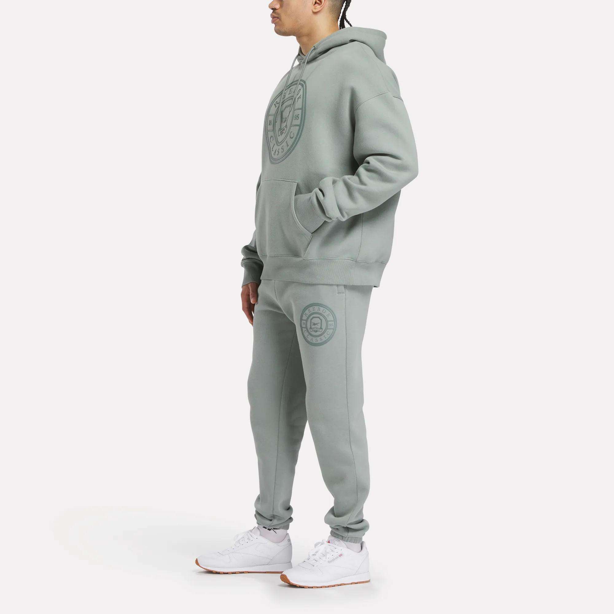A man in Reebok Men's Crest Jogger Pants and a matching light green hooded sweatshirt with logos, paired with white sneakers, stands against a plain white background, highlighting laid-back style and comfort.