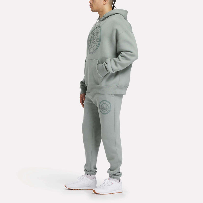 A man in Reebok Men's Crest Jogger Pants and a matching light green hooded sweatshirt with logos, paired with white sneakers, stands against a plain white background, highlighting laid-back style and comfort.