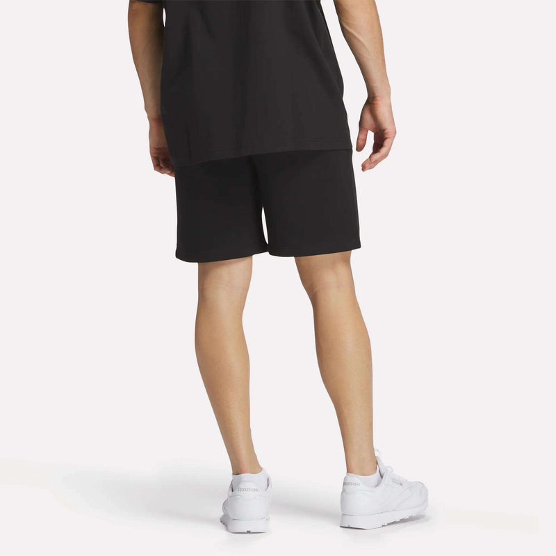 A person stands with their back turned, showing only the lower half, wearing Reebok Men's Crest Shorts in black cotton and white sneakers against a plain light background.