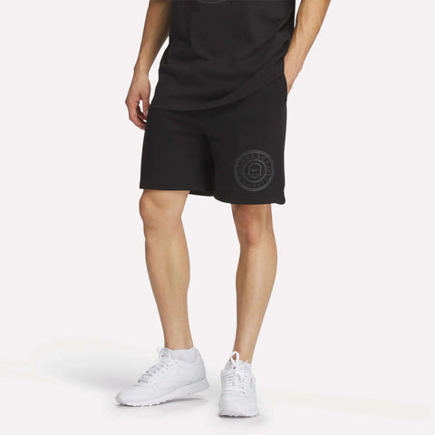 A person in a black t-shirt and Reebok Men's Crest Shorts with a round logo on the left leg, paired with white sneakers, stands against a plain white background.
