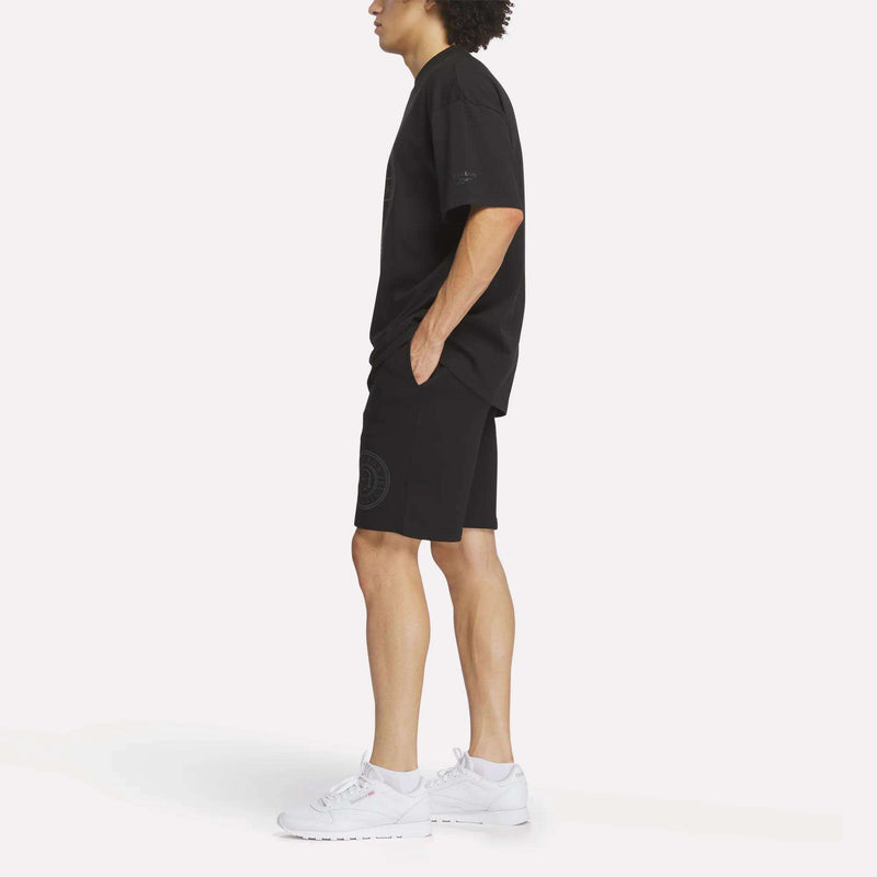 A person stands in profile wearing a loose black T-shirt, Reebok Men's Crest Shorts in a relaxed fit, and white sneakers against a plain white background.