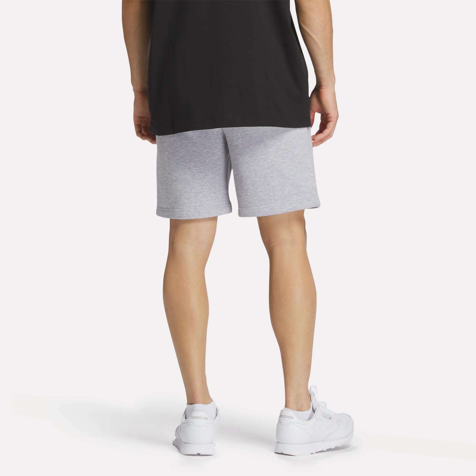 A person wearing a black T-shirt, Reebok Men's Crest Shorts in light gray (relaxed fit), and white sneakers is shown standing with their back to the camera against a plain white background.