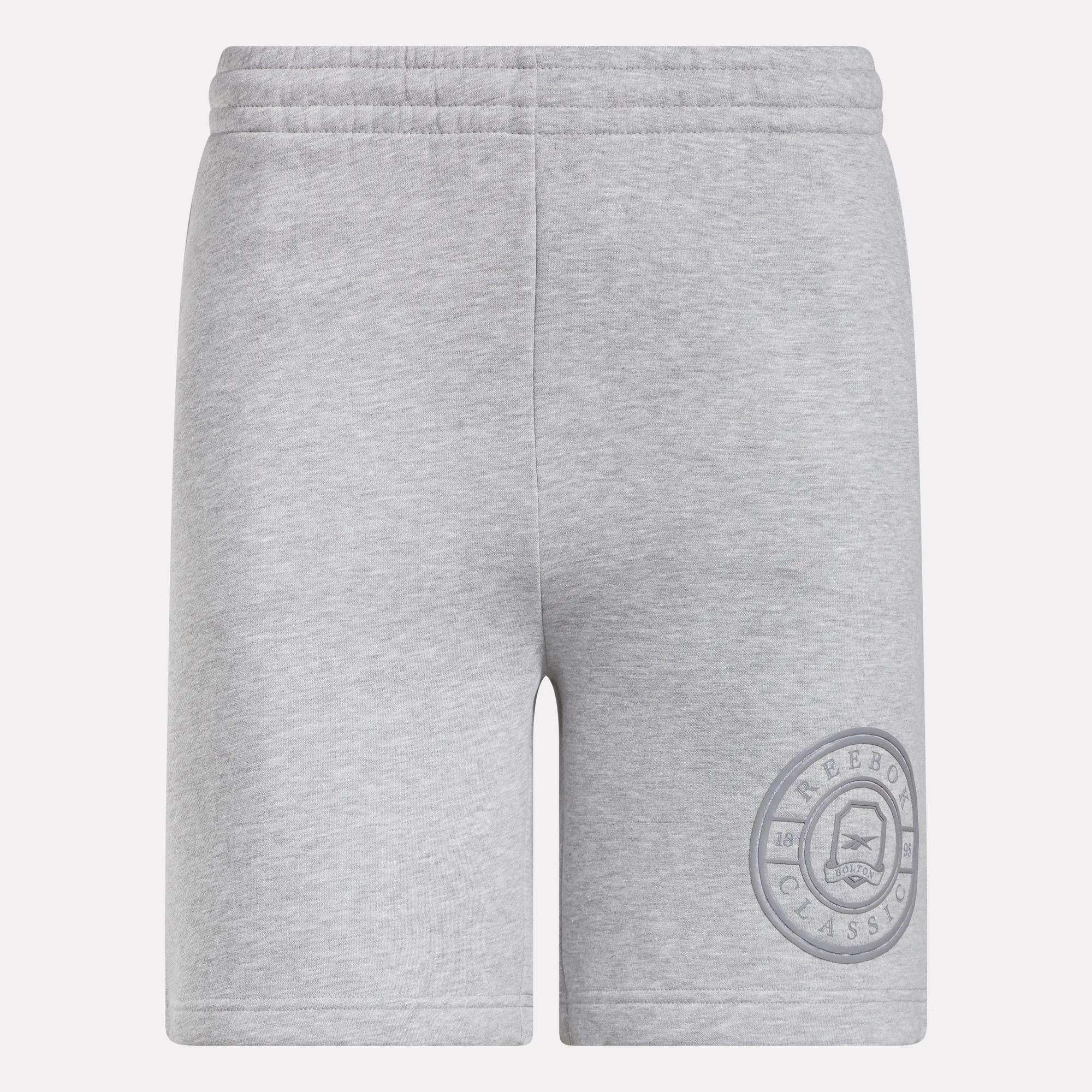 Reebok Men's Crest Shorts in light grey feature an elastic waistband and relaxed fit. Made from cotton, these shorts have a simple design without visible pockets or drawstrings and display a circular Reebok Classic logo on the lower left leg.