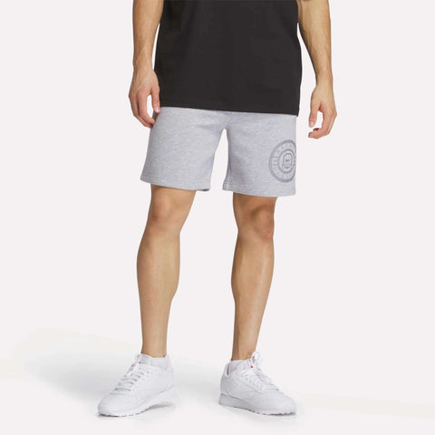 A person in a black T-shirt, Men's Reebok Crest Shorts featuring the logo on the left leg, and white sneakers stands against a plain white background.