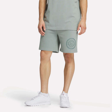 A person wears Reebok Men's Crest Shorts—relaxed fit, light green cotton with a circular logo on the left leg—and a matching tee with white sneakers, standing against a plain white background.