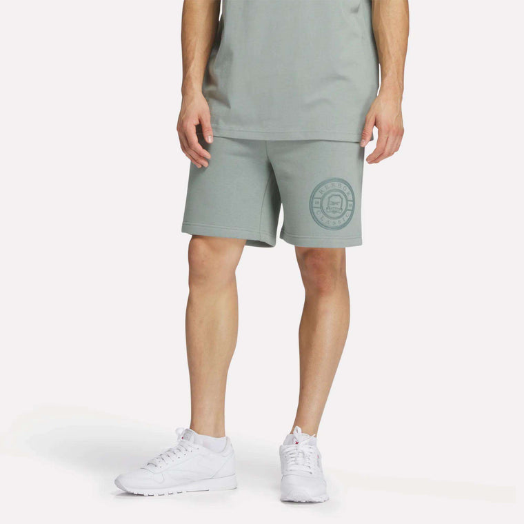 A person wears Reebok Men's Crest Shorts—relaxed fit, light green cotton with a circular logo on the left leg—and a matching tee with white sneakers, standing against a plain white background.
