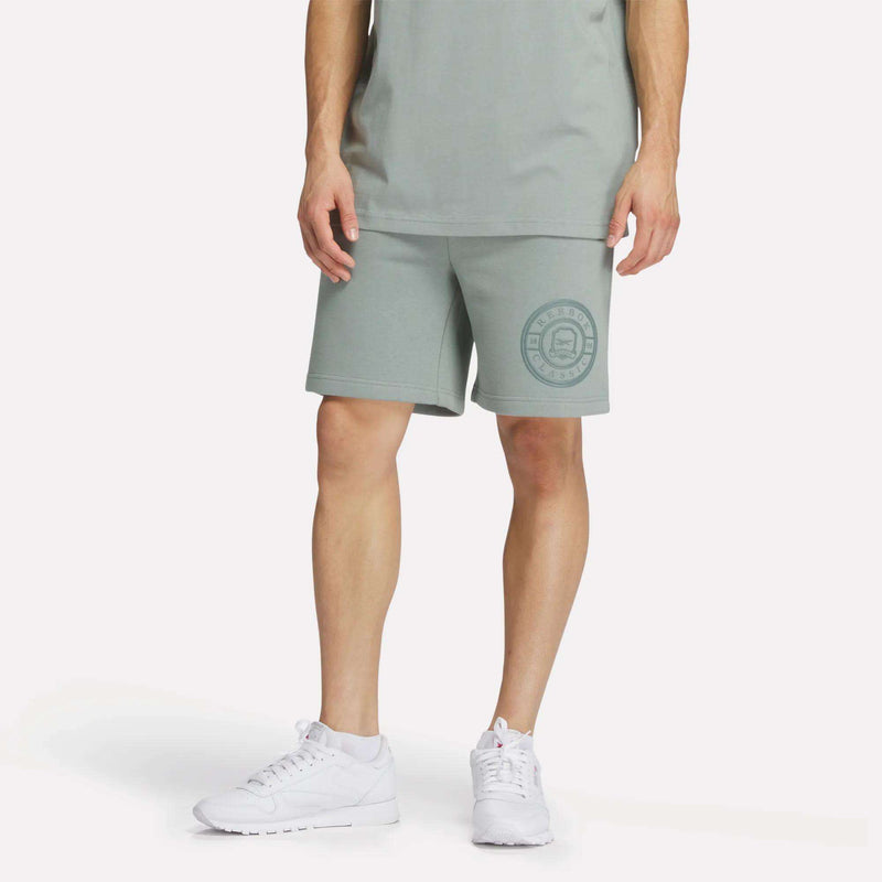 A person wears Reebok Men's Crest Shorts—relaxed fit, light green cotton with a circular logo on the left leg—and a matching tee with white sneakers, standing against a plain white background.