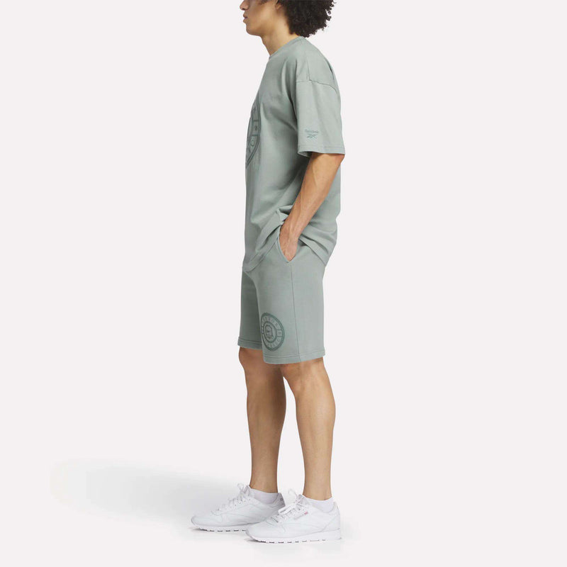 A person stands in profile against a white background, hands in their pockets, wearing a relaxed fit light sage green T-shirt and matching Reebok Men's Crest Shorts paired with white sneakers.