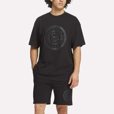 A person wears a black oversized Reebok Men's Crest T-Shirt and matching black shorts with Reebok logos, standing against a plain light background—an ideal choice for everyday comfort.