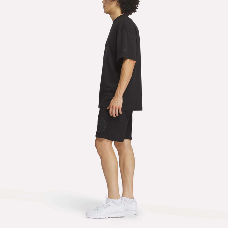 A person stands sideways in a Reebok Men's Crest T-Shirt, paired with black shorts and white sneakers, highlighting everyday comfort against a plain white background.
