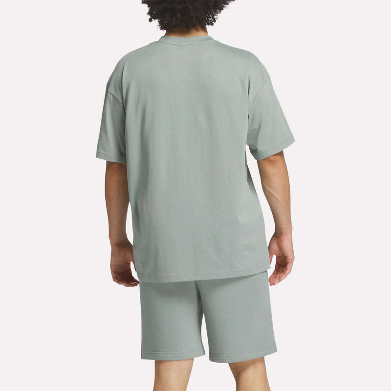 A person with curly hair stands with their back to the camera, wearing a loose Men's Crest T-Shirt by Reebok and matching shorts. The plain light background highlights this comfortable, everyday style.