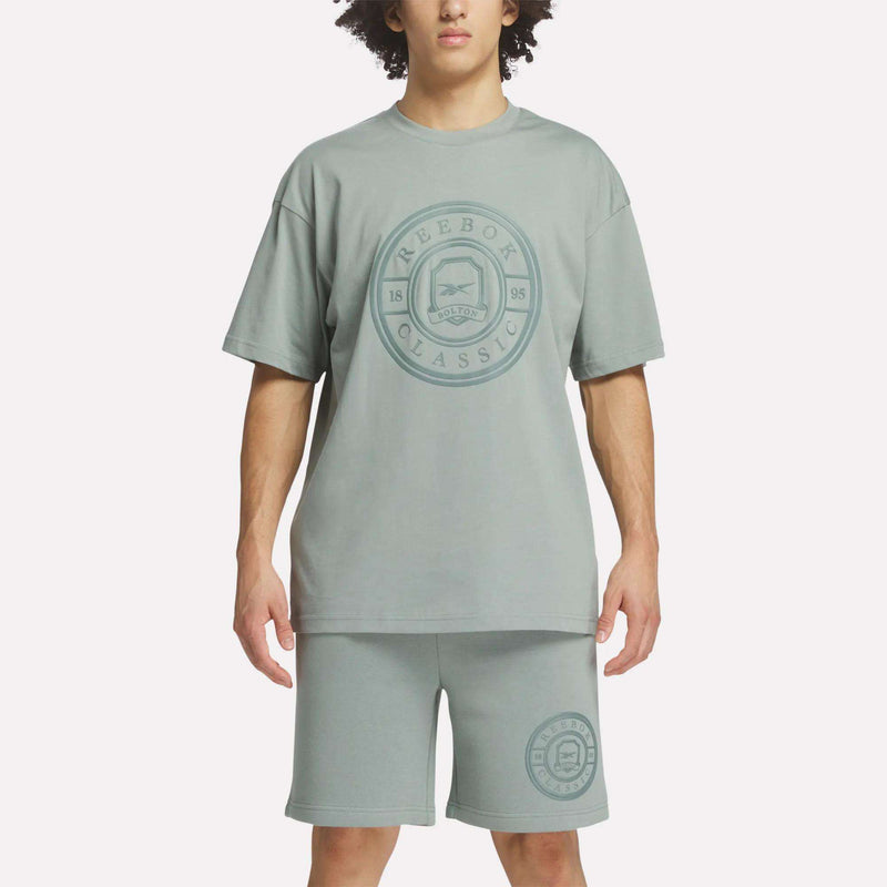 A person wears a loose-fitting, light green Reebok Men's Crest T-Shirt and matching shorts with large circular logos. The cotton tee provides everyday comfort, shown against a plain white background.