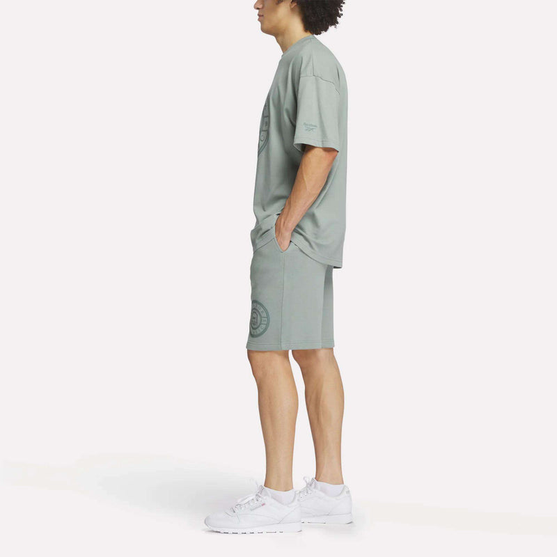 A man stands in profile against a white background, hands in his pockets, wearing the Reebok Men's Crest T-Shirt with matching shorts and white sneakers for a relaxed, everyday style.