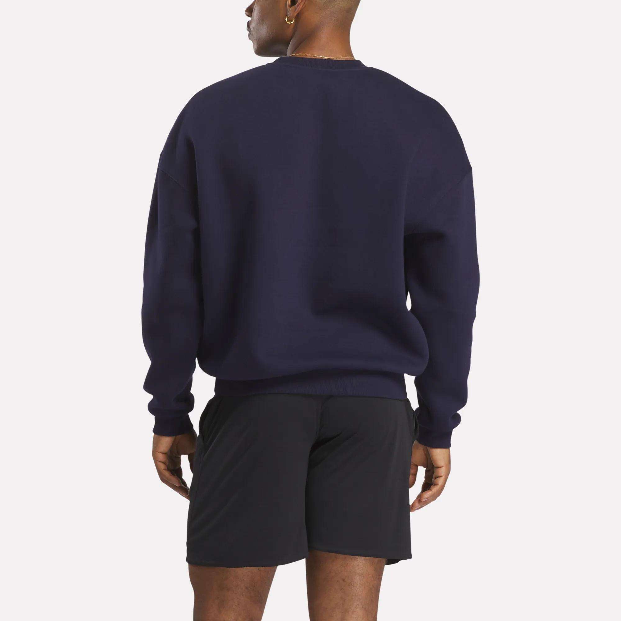 A person stands with their back to the camera, wearing a Reebok Men's Crew Sweatshirt in dark navy and black shorts, against a plain white background.