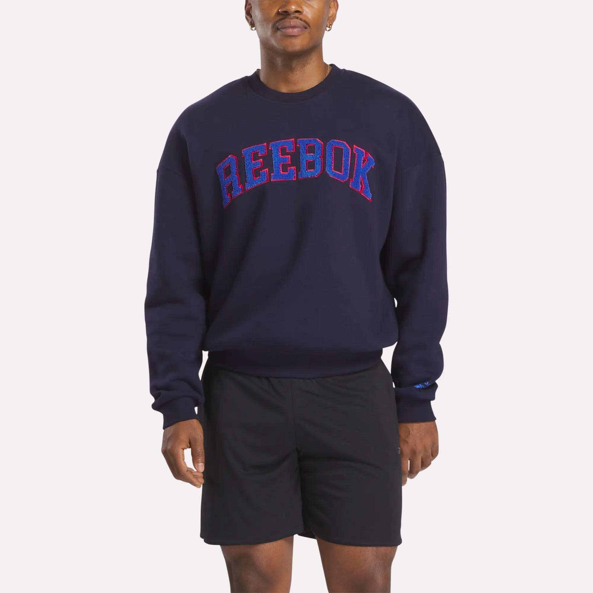A man stands against a plain background, wearing a Reebok Men's Crew Sweatshirt in navy blue with bold red and blue lettering, paired with black athletic shorts. His arms are relaxed by his sides.
