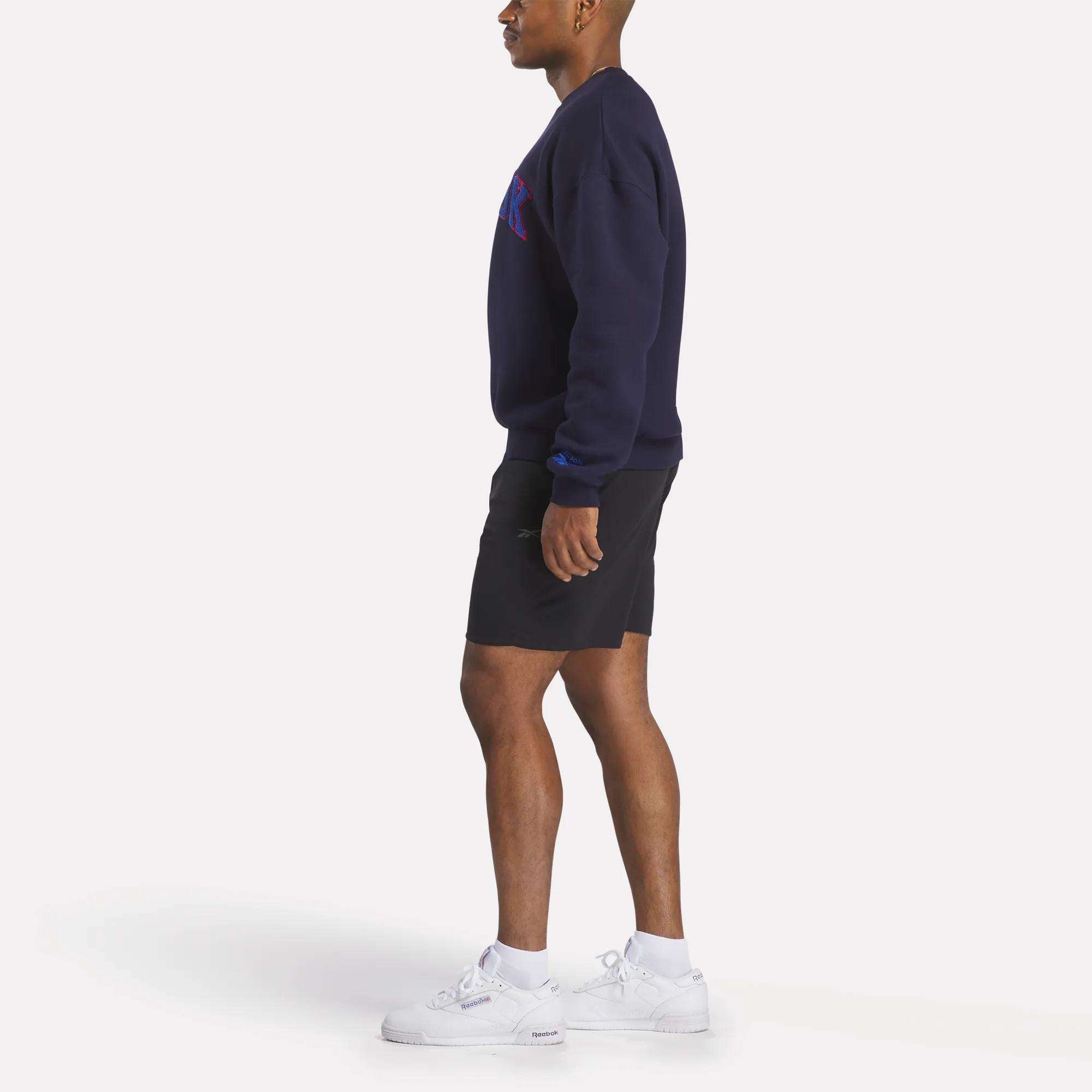 A person in profile wears a Reebok Men's Crew Sweatshirt in navy, paired with black shorts, white socks, and white sneakers against a plain white background.