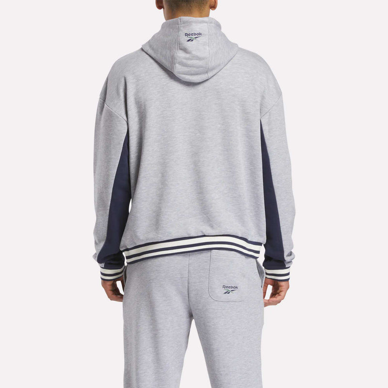 A person wearing a light gray Reebok Men's Lawn Club French Terry Hoodie, seen from behind. This hoodie features dark blue sleeve accents and striped cuffs and waistband.