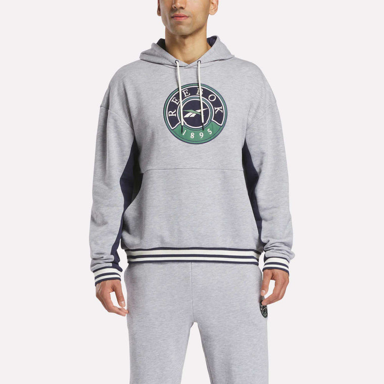 A man stands against a plain white background wearing the Reebok Men's Lawn Club French Terry Hoodie in light gray with a green and navy logo, paired with matching sweatpants. This cotton blend hoodie offers comfort and style.