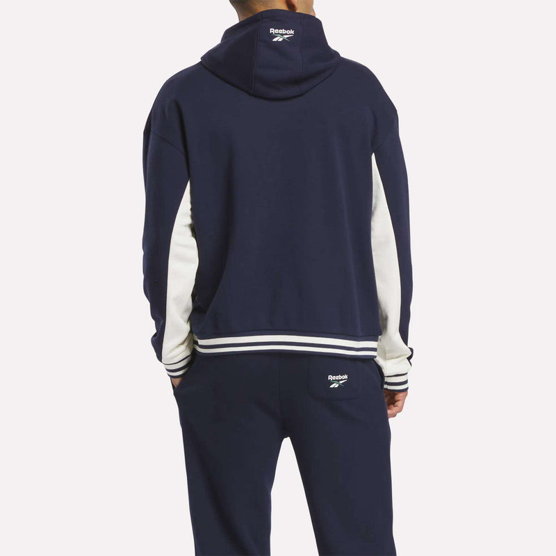 A person faces away wearing the Reebok Men's Lawn Club French Terry Hoodie tracksuit in navy blue with white sleeve stripes and trim. The cotton-blend hoodie displays the Reebok logo on the hood's back and pants pocket.