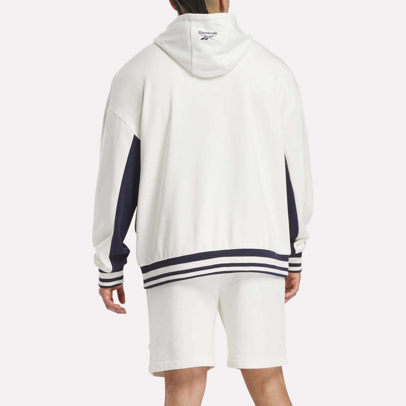 A person stands facing away, wearing the Reebok Men's Lawn Club French Terry Hoodie in white with navy stripes on the hem and cuffs, plus a visible Reebok logo on the hood, paired with white shorts.