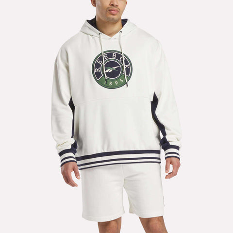A person wears a white Reebok Men's Lawn Club French Terry Hoodie with a green and navy logo, navy stripes on cuffs and hem, and matching white shorts, standing against a plain background.