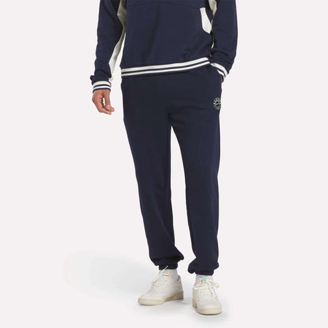A person wears Reebok Men's Lawn Club French Terry Jogger Pants in navy with white details and white sneakers, standing with one hand in a pocket against a plain light background, shown from shoulders down.