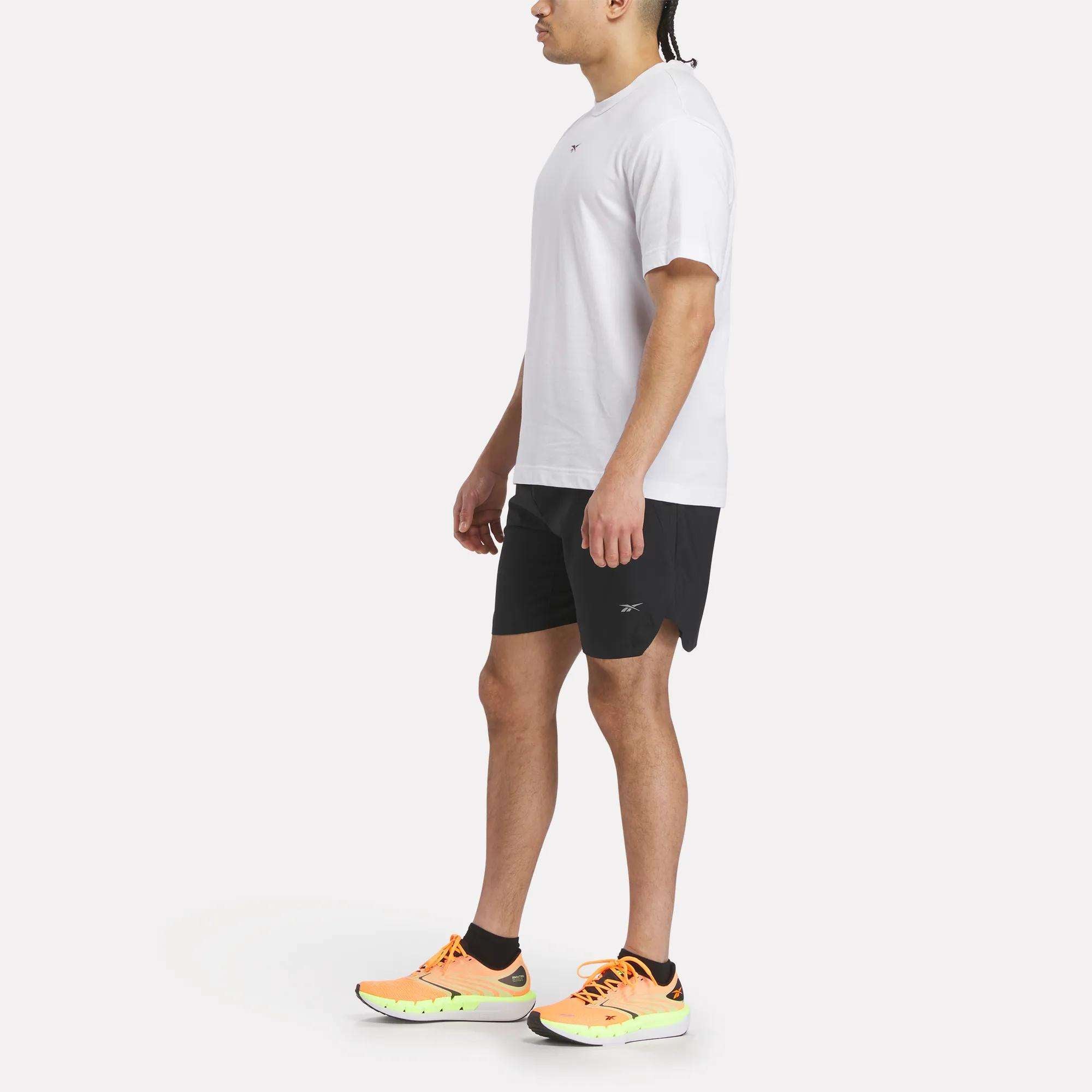 A person stands sideways wearing a white Reebok Men's Athlete Speed Logo T-Shirt, black shorts, black socks, and bright orange athletic shoes with yellow soles against a plain light background.