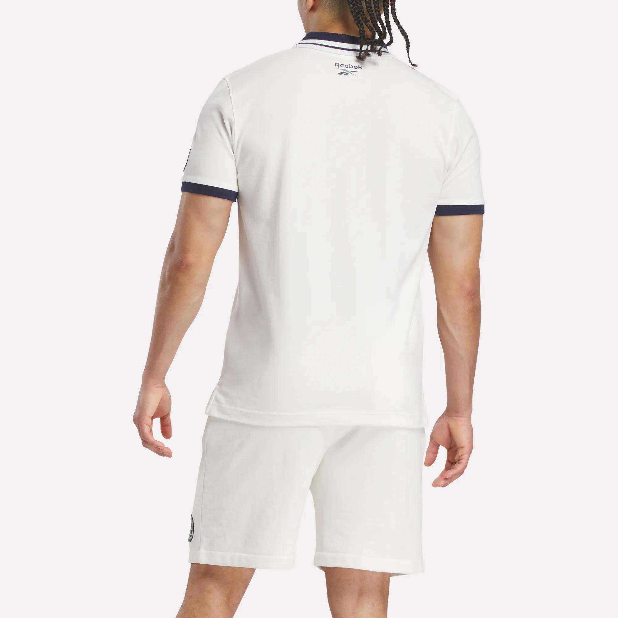 A man with braided hair stands with his back to the camera, wearing a white Reebok Men's Lawn Club Polo Shirt and white shorts with dark trim. The comfortable cotton-blend polo fits well. The background is plain and light-colored.