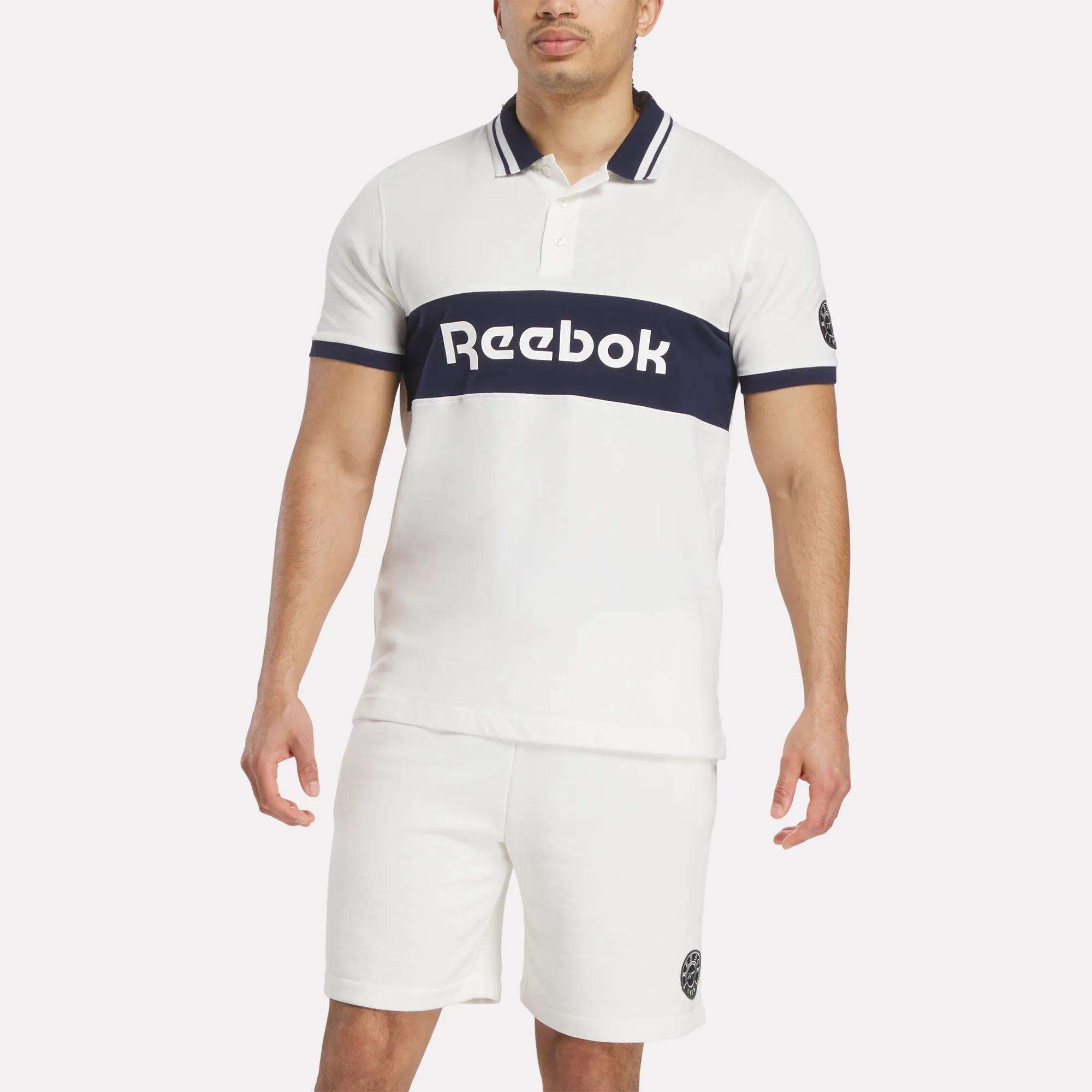 A man wears a white Reebok Men's Lawn Club Polo Shirt with a navy stripe and logo, paired with matching white shorts featuring a small Lawn Club logo on the left leg.