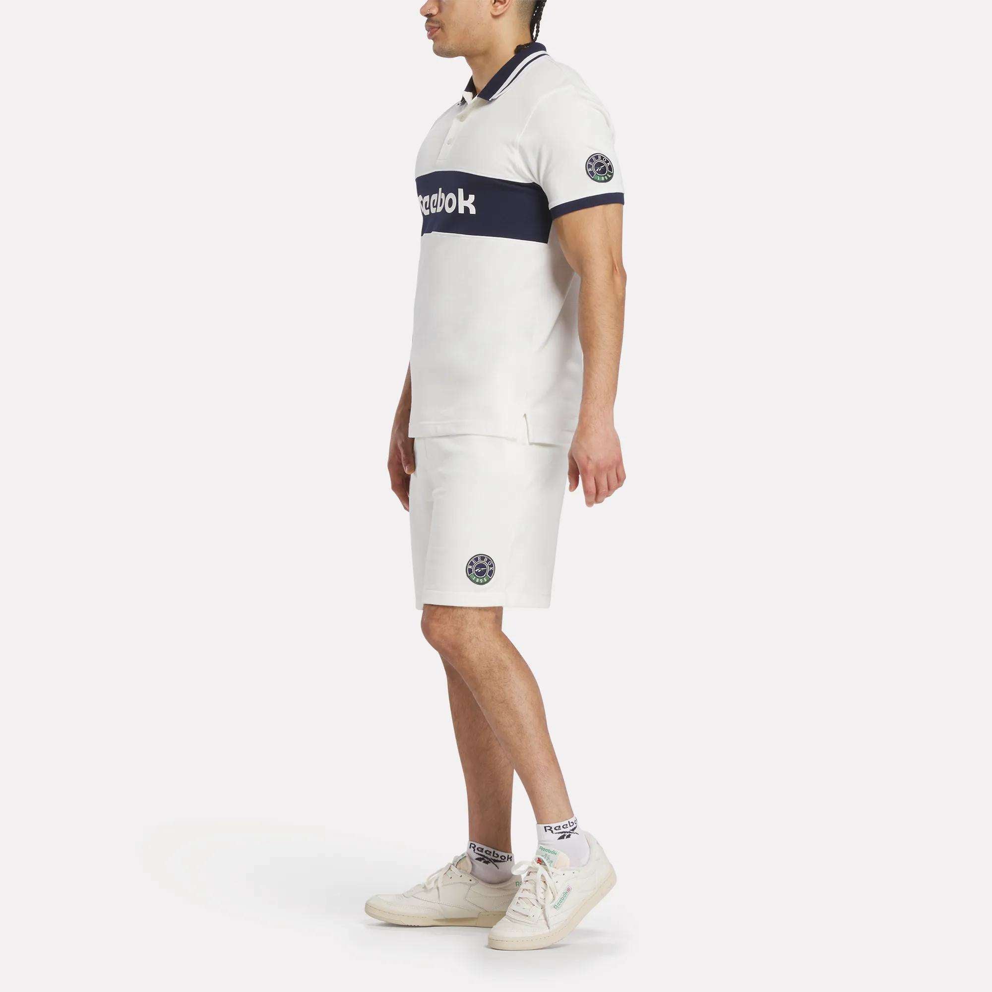 A man stands in profile wearing a Reebok Men's Lawn Club Polo Shirt with navy details, paired with matching white shorts, white and blue-accented socks, and white sneakers against a plain light background.