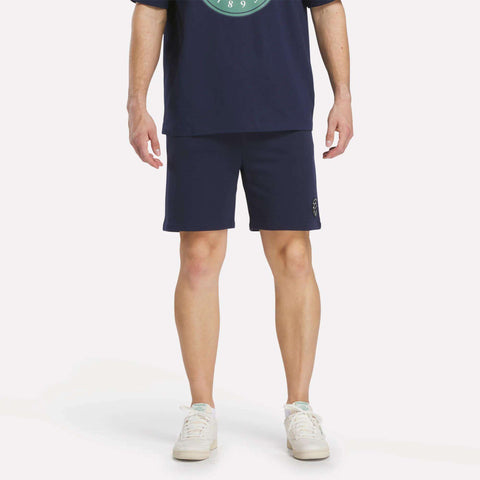 A person stands with arms relaxed by their sides, wearing a navy blue T-shirt, Reebok Men's Lawn Club Shorts, and white sneakers. The image is cropped at the shoulders, so their face is not visible.
