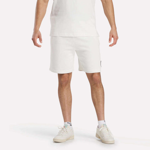 A person wearing a white t-shirt, Reebok Men's Lawn Club Shorts in white, and white sneakers stands against a plain white background, shown from shoulders to feet.