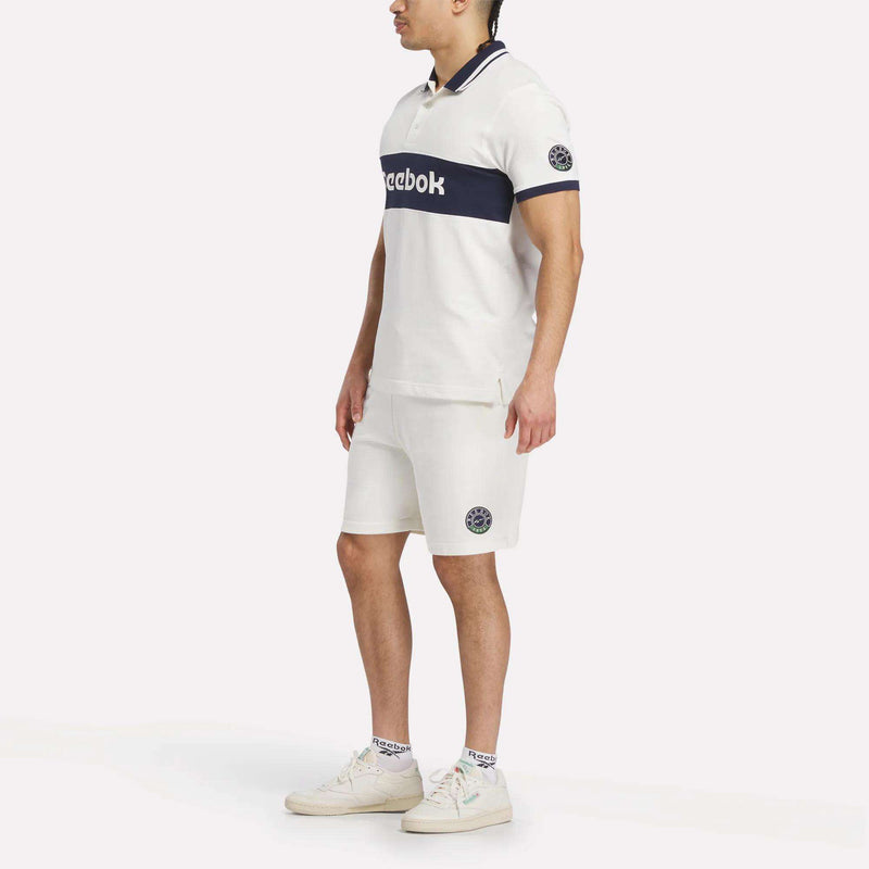 A man stands on a plain light background wearing Reebok’s Men's Lawn Club Shorts with navy blue accents, a matching white Reebok polo shirt, white Reebok sneakers, and white socks—all featuring the iconic Reebok logo.
