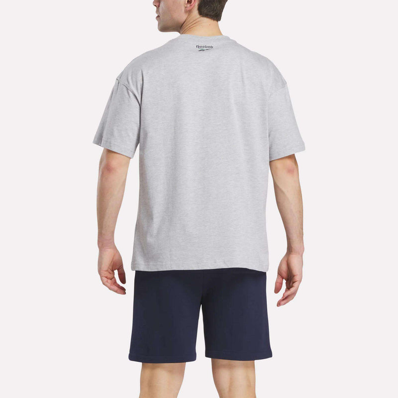 A man is seen from the back wearing a light gray Reebok Men's Lawn Club T-Shirt and navy blue shorts against a plain, light background.