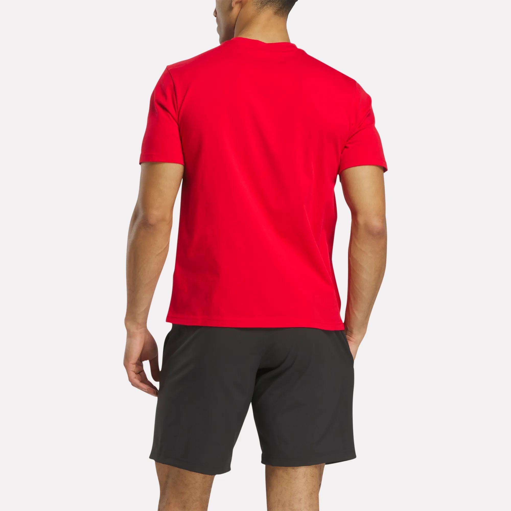 A person stands facing away against a plain white background, wearing a bright red Reebok Men's Collegiate T-Shirt and black shorts.