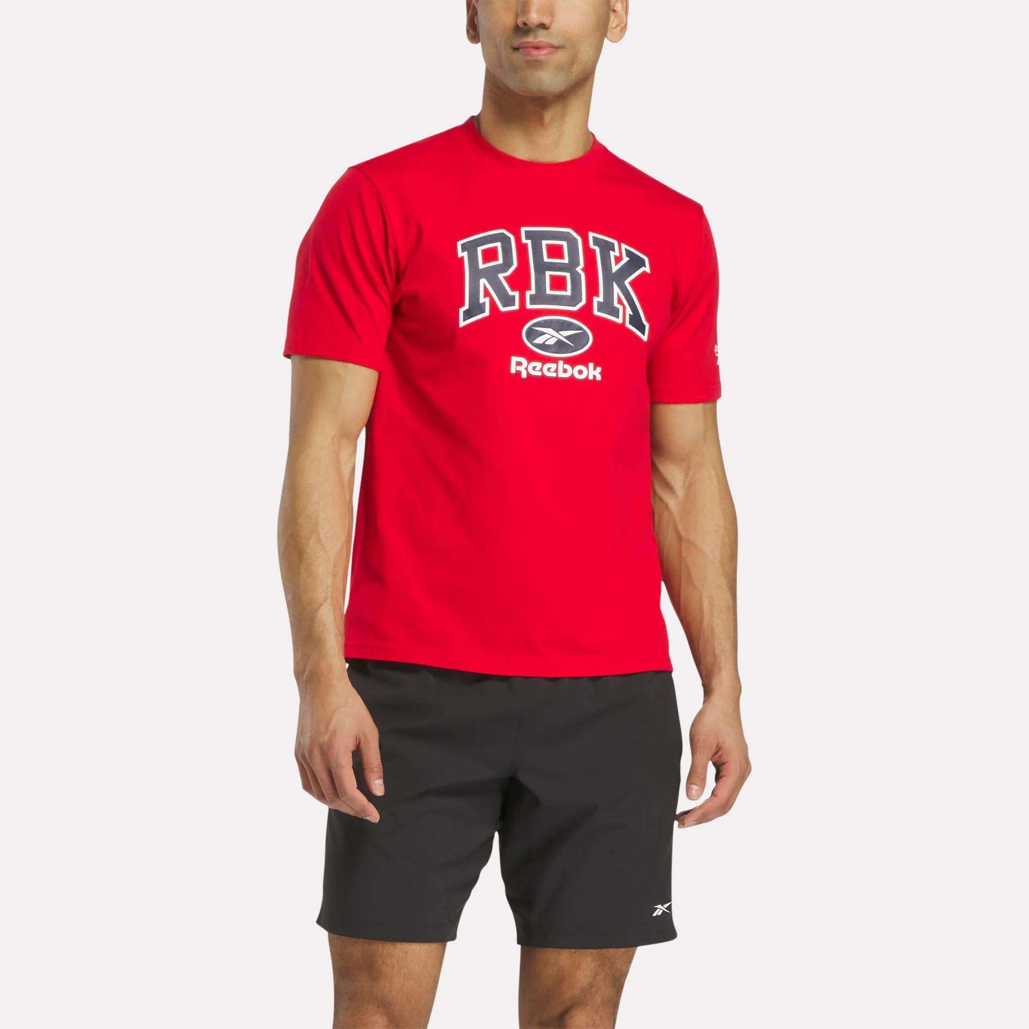 A man wears a Reebok Men's Collegiate T-Shirt—bright red with RBK in white and black—paired with black athletic shorts, standing against a plain white background. The casual crewneck is made from comfortable cotton.