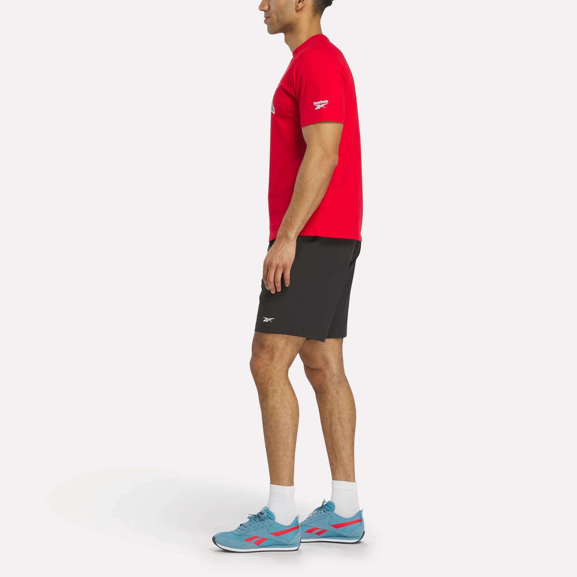 A man standing sideways wears a red Reebok Men's Collegiate T-Shirt, black shorts, white socks, and blue athletic shoes against a plain white background.
