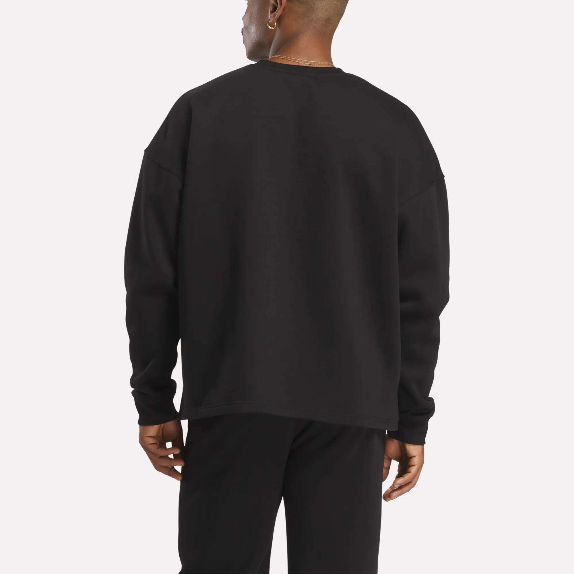 Facing away from the camera against a plain, light background, a person wears Reebok's Men's Wardrobe Essentials Sweatshirt with black pants, featuring a relaxed fit and crafted from soft cotton-blend fleece.