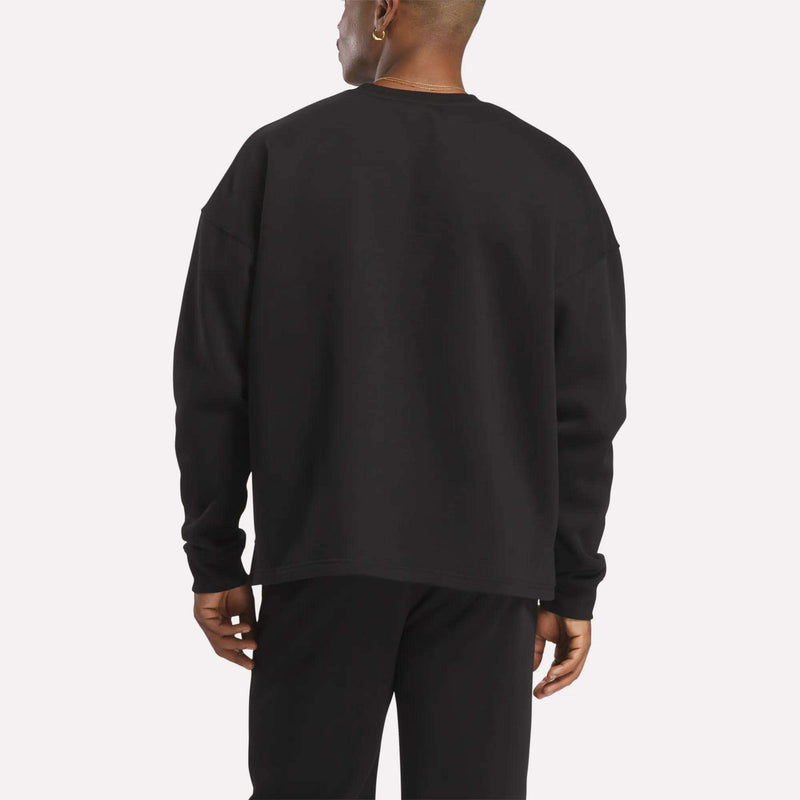 Facing away from the camera against a plain, light background, a person wears Reebok's Men's Wardrobe Essentials Sweatshirt with black pants, featuring a relaxed fit and crafted from soft cotton-blend fleece.