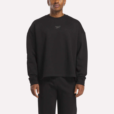 A man wears the Reebok Men's Wardrobe Essentials Sweatshirt, a relaxed fit in soft cotton-blend fleece with a small white chest logo, styled with black pants as he stands facing forward against a plain background.