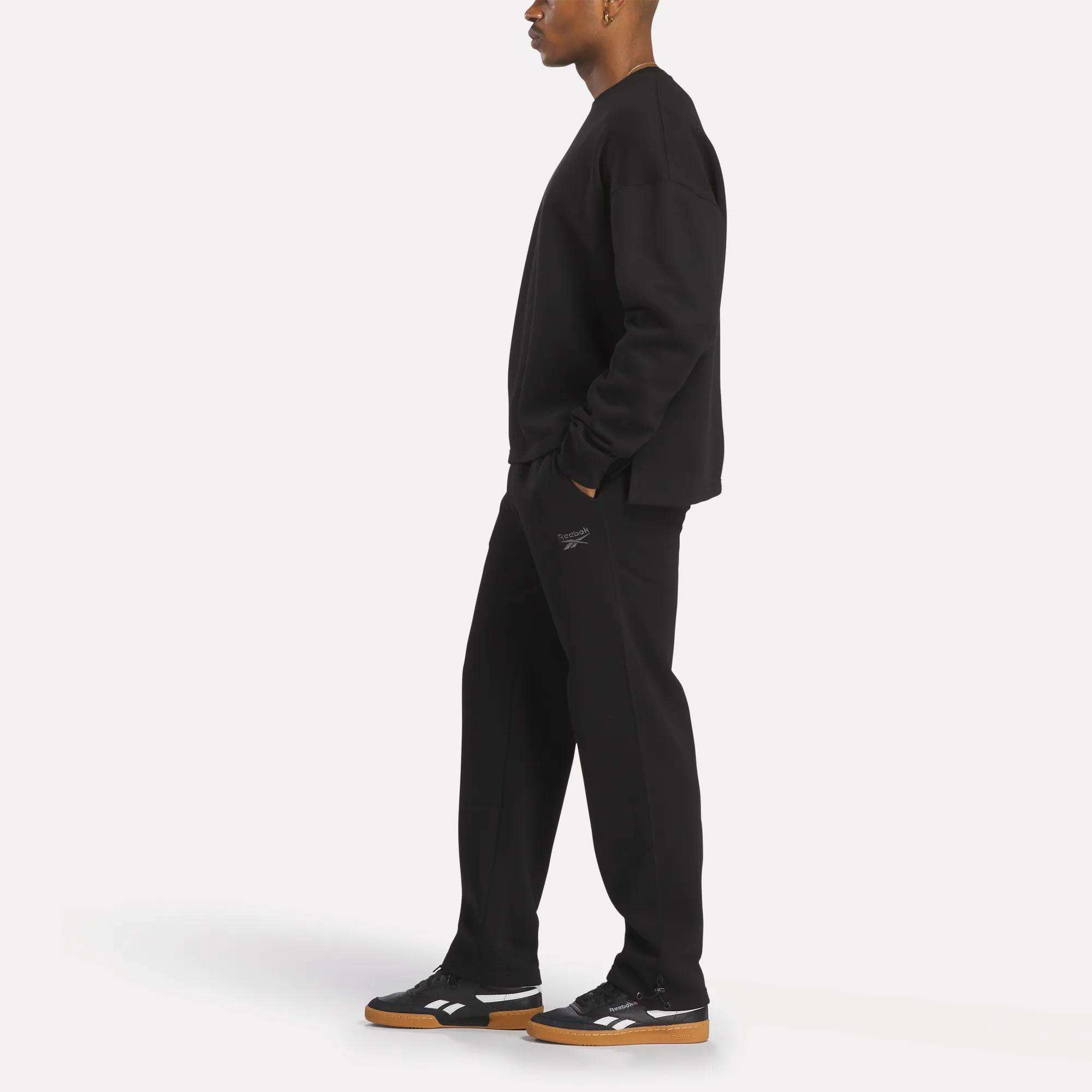 A person in profile wears a relaxed fit Men's Wardrobe Essentials Sweatshirt by Reebok and black pants with a small thigh logo. Their black Reebok sneakers have white accents and gum soles, set against a plain white background.