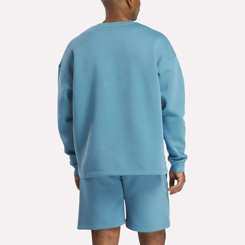 A person faces away from the camera, wearing a relaxed-fit light blue Reebok Men's Wardrobe Essentials Sweatshirt and matching cotton-blend fleece shorts, set against a plain white background.
