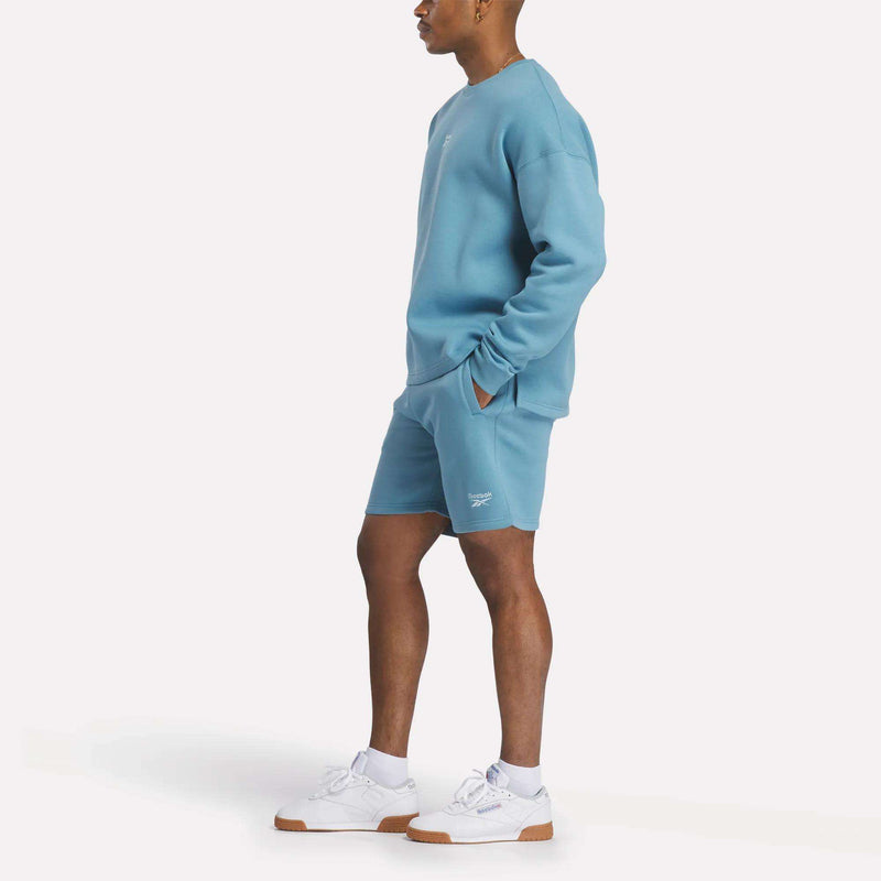 A person stands sideways wearing the Reebok Men's Wardrobe Essentials Sweatshirt and matching shorts in a relaxed fit with white sneakers. Their hands are in their pockets against a plain white background.